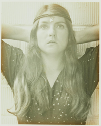 Image of Bea Nettles using the alchemical method of being a gelatin silver print. Shows Bea wearing a star-patterned button up shirt, her hair is down and Bea has a headband of two rope-like strings wrapping around her head with a star dangling from the center onto her forehead. She is looking to the front with eyes wide open, she has her arms pulled back with her hands behind her head.