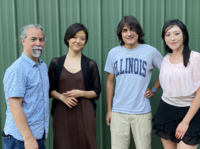 Photo of Kuroshio members all smiling at the camera: Jason Finkelmen (dressed in a blue speckled button-up with dark blue jeans), Saori Kataoka (dressed in a brown long dress with a black cardigan), Kavi Naidu (dressed in a grey Illinois t-shirt with pale khaki cargo pants), Joy Yang (dressed in a pale pink blouse and a black skirt).