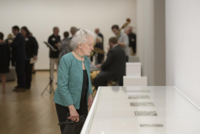 Image of 2019 Acquired Taste Celebration, there can be seen a participant in all black with a teal jacked and her hair pulled back while looking at a display in front of here; in the background there is a blurred audience of which is comprised of other participants viewing art as well as musicians playing the bass, keyboard, and drums.