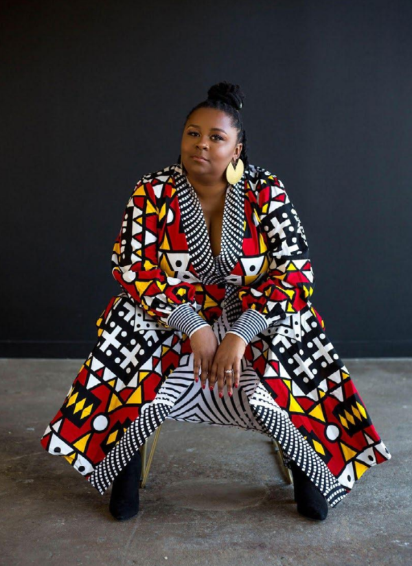 Image of Founder, President, and CEO of Creative Reaction Lab, Antoinette Carroll sitting in a chair with her body forward, putting her weight onto her elbows. She wears a patterned outfit in white, black, red, and yellow with black boots.