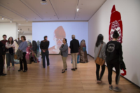 Image of School of Art + Design Master of Fine Arts Exhibition installation at Krannert Art Museum from 2018. There can be seen guests taking the art projected on the wall as well as the red art installed on the right wall.