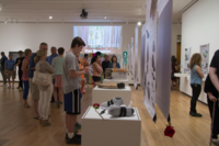 Image of School of Art + Design Bachelor of Fine Arts Exhibition, installation at Krannert Art Museum from 2018. There can be seen various parents, students, and young kids admiring items on display such as shoes beside a rose.