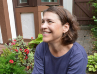 Image of speaker Dede Fairchild Ruggles from an angle. She is smiling a tooth smile. There can be seen red flowers and green leafy plants in the background.