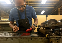 Artist and printmaker Ben Blount, a black man with glasses, a blue T shirt, and a black apron, stands at a press, holding a red roller filled with ink. He is in his studio, an industrial space with other press equipment.