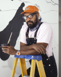 A grown man with a beard and mustache wearing an orange baseball cap, black glasses, a blue bandana around his neck, and a white t-shirt with black overalls leaning over a ladder with a cup of paintbrushes in his left hand.
