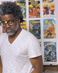 Middle aged man wearing a white v-neck t-shirt with black rimmed roundish glasses with tape wrapped around the nose piece posing in a heroic stance in front of a wall decorated with comic books.