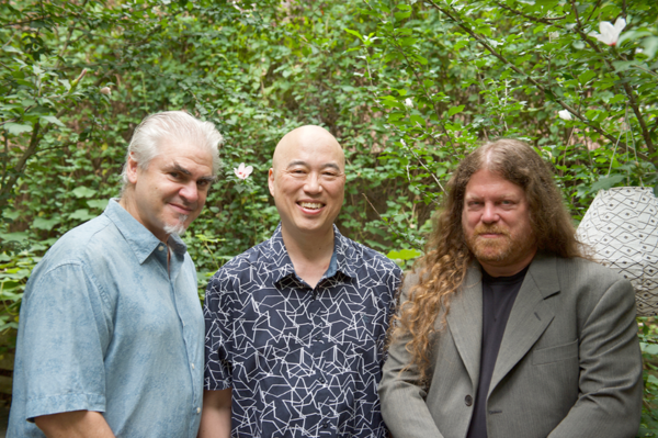 Three adult men looking ahead and smiling in front of green leafed trees.