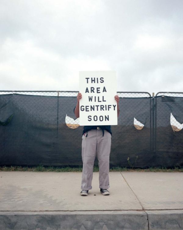 person holding a white sign in front of their face and torso that reads "this area will gentrify soon". Background is a wired fence with a black tarp strewn about the fence.