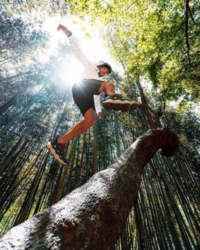 adult male model jumping onto a tree in the middle of a green forest, displaying outdoor gear and footwear