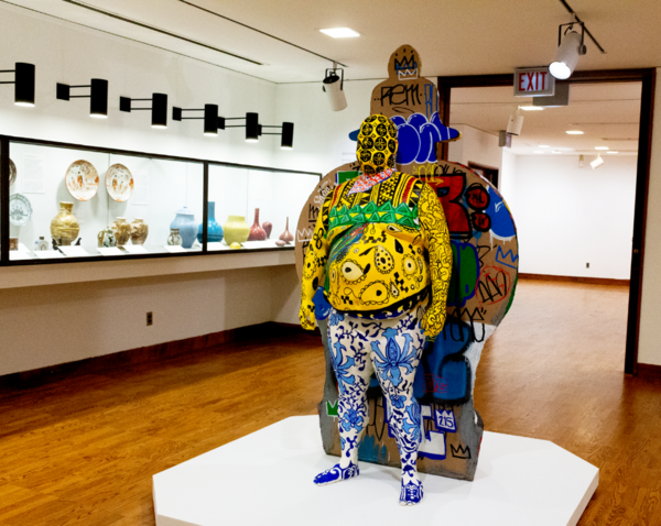 painted statue standing tall in front of a cardboard with designs. The statue is decorated in vibrant colors and patterns and the half jar designed cardboard behind the statue is full of graffiti tags