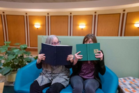 Liz Stine-Morrow and Melissa Troyer sitting, side-by-side, on a sofa at the Beckman Institute, mischievously holding open books in front of their faces to shield all but their eyes.